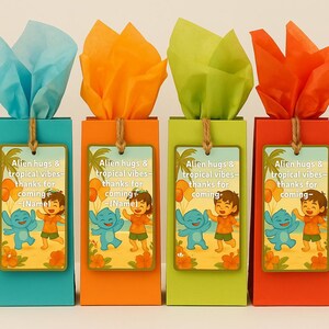 May include: Four colorful gift boxes with tissue paper and tags. The boxes are teal, orange, green, and red. Each box has a tag with the text "Alien hugs & tropical vibes - thanks for coming - (Name)" and a cartoon illustration.