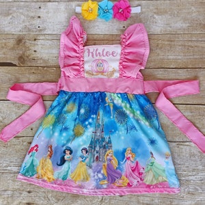May include: A pink princess dress with a blue and white print featuring Cinderella's castle, fireworks, and Disney princesses. The dress has a pink sash and a white bodice with the name "Khloe" embroidered on it. The dress is accompanied by a headband with three flowers in yellow, blue, and pink.
