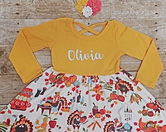 May include: Mustard yellow long-sleeved dress with a white skirt featuring a Thanksgiving-themed print. The dress has a criss-cross back and the name "Olivia" in white script. A matching floral headband completes the outfit.