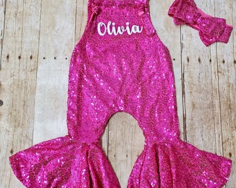 May include: Pink sequined overalls with a bell bottom style and the name "Olivia" embroidered on the front. A matching pink sequined headband is included.