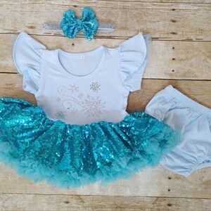 May include: A light blue baby outfit set featuring a short-sleeved top with snowflake design, a sequined turquoise tutu skirt, matching bloomers, and a sequined bow headband. The set is designed for infants.