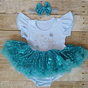 May include: A light blue baby outfit with a sequined turquoise tutu skirt and matching bow headband. The top has flutter sleeves and a snowflake design. The outfit is designed for infants and toddlers.