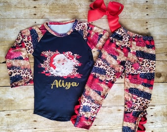 May include: A Christmas-themed pajama set featuring a navy blue long-sleeved top with a Santa Claus graphic and the name "Aliya" in gold. The sleeves have a leopard print pattern. The matching pants and a red bow complete the set.