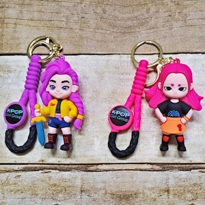May include: Three keychains featuring cartoon figures with gold-colored rings. Each keychain has a figure with a unique outfit and hairstyle. The keychains have a black and colored strap with the text "K-POP Girl Group" on a circular tag.