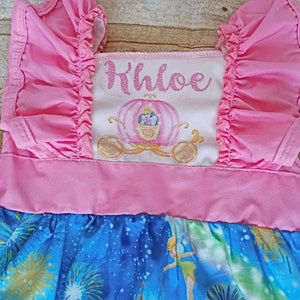 May include: Pink and blue dress with a white panel featuring a Cinderella carriage and the name Khloe in pink glitter. The dress has a ruffled bodice and a blue skirt with a Tinkerbell and fireworks print.