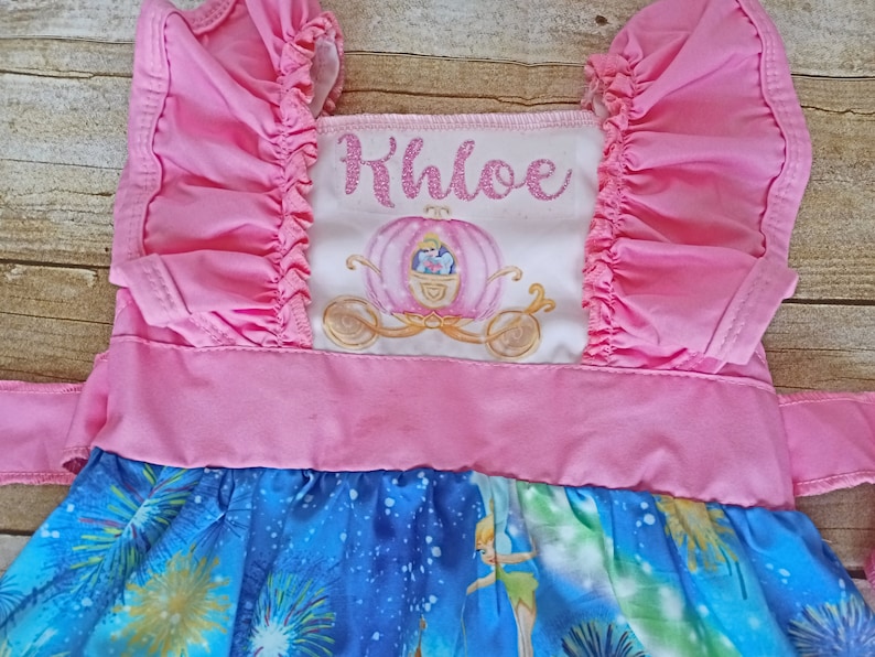May include: Pink and blue dress with a white panel featuring a Cinderella carriage and the name Khloe in pink glitter. The dress has a ruffled bodice and a blue skirt with a Tinkerbell and fireworks print.