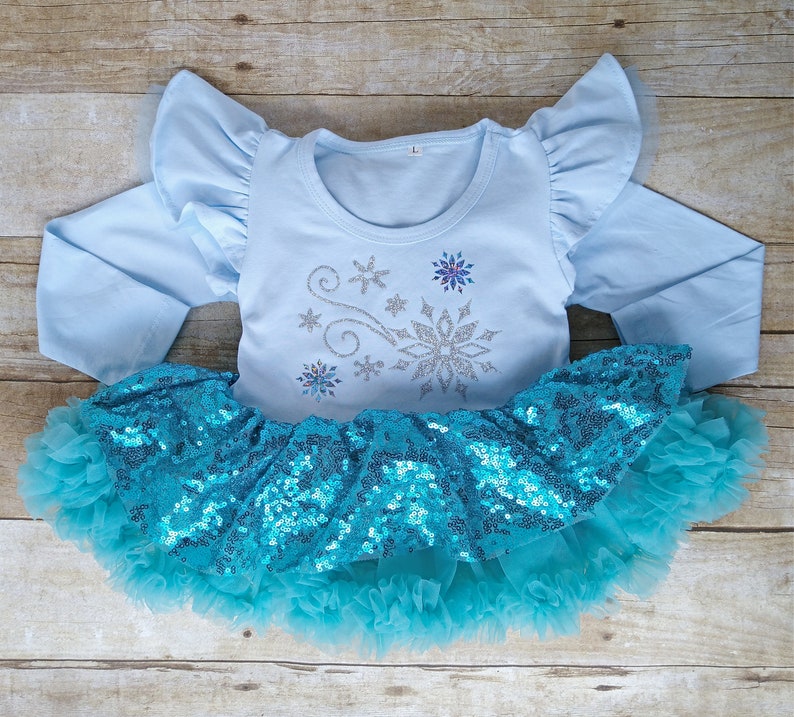 May include: A light blue long-sleeved top with ruffled sleeves and a matching turquoise sequined tutu skirt. The top features a silver snowflake design with glitter accents. The skirt has a layered tulle hem.