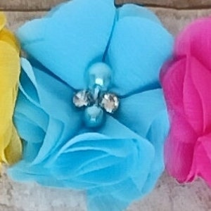 May include: A white headband with three fabric flowers. The flowers are yellow, blue, and pink. Each flower has a pearl and rhinestone embellishment in the centre.