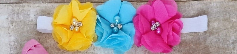 May include: A white headband with three fabric flowers. The flowers are yellow, blue, and pink. Each flower has a pearl and rhinestone embellishment in the centre.