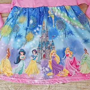 May include: A pink dress with a blue and purple print featuring Disney princesses and Cinderella's castle. The print also includes fireworks and stars.