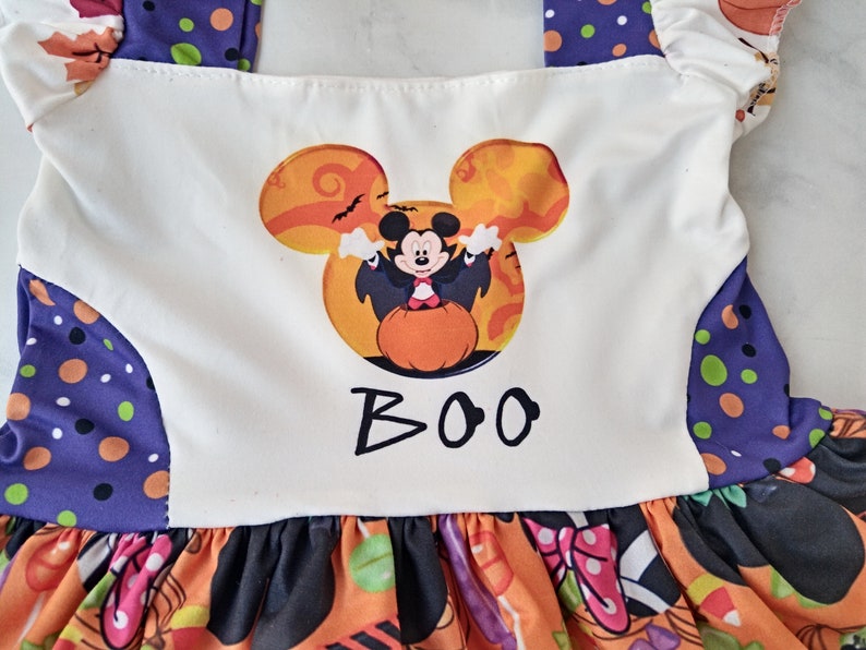 May include: A Halloween dress with a Mickey Mouse design. The white bodice features a Mickey Mouse head graphic in orange and black, with the word "BOO" underneath. The skirt is orange with Halloween patterns.