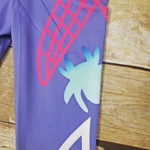 May include: Purple leggings with a pink and white geometric design. The design features a large pink and white geometric shape, a smaller blue and white geometric shape, and a white geometric shape with a pink outline.