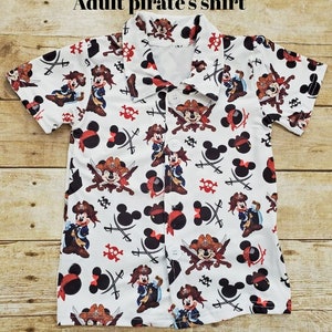 May include: White short-sleeved button-up shirt with a repeating pattern of Mickey Mouse dressed as a pirate. The shirt has a white background with red, black, and brown accents.