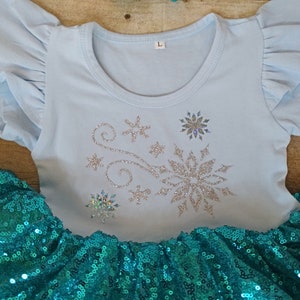 May include: A light blue toddler's outfit featuring a top with silver glitter snowflake designs and ruffled sleeves. The outfit includes a teal sequined skirt. The top has the letter "L" printed on the collar.