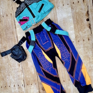 May include: A two-piece outfit featuring a turquoise crop top with black accents and a matching pair of blue and black pants with orange and floral details. A small black bag with a strap is also included. The outfit is displayed on a wooden surface.