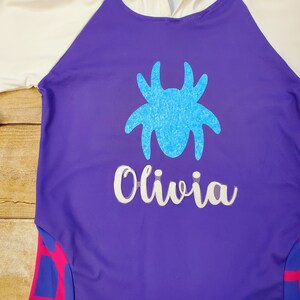 May include: A purple long-sleeved shirt with a white and pink striped design on the sleeves. The shirt has a blue glitter spider and the name "Olivia" in white glitter on the front.