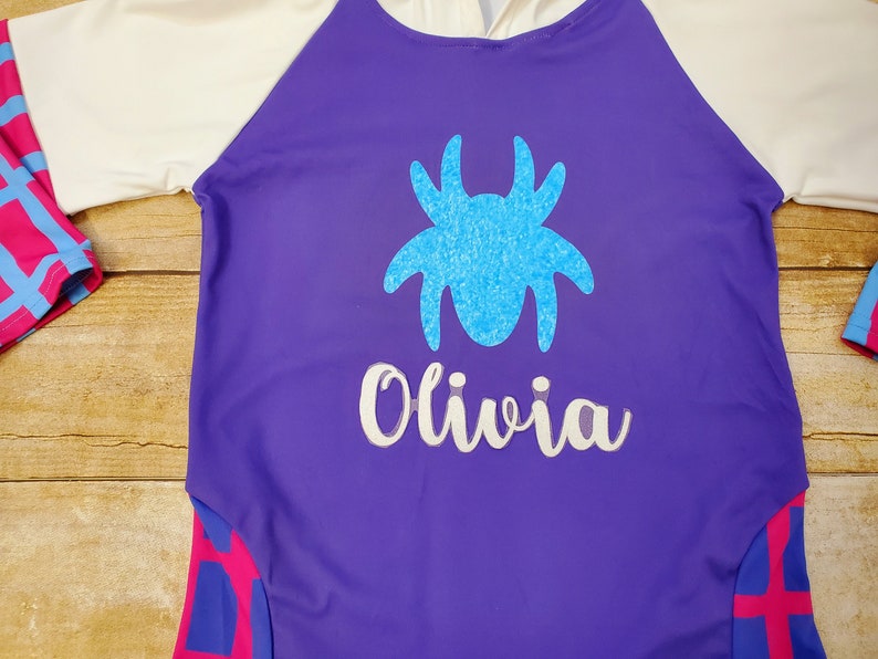 May include: A purple long-sleeved shirt with a white and pink striped design on the sleeves. The shirt has a blue glitter spider and the name "Olivia" in white glitter on the front.