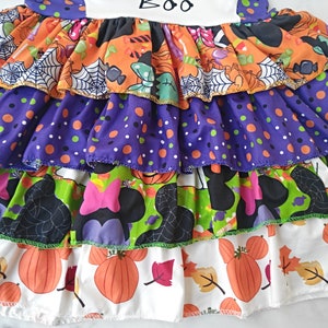 May include: A multi-layered Halloween dress with a variety of patterns including pumpkins, polka dots, spiderwebs, and Minnie Mouse silhouettes. The top layer has the word "BOO".