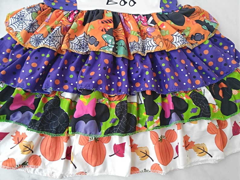 May include: A multi-layered Halloween dress with a variety of patterns including pumpkins, polka dots, spiderwebs, and Minnie Mouse silhouettes. The top layer has the word "BOO".