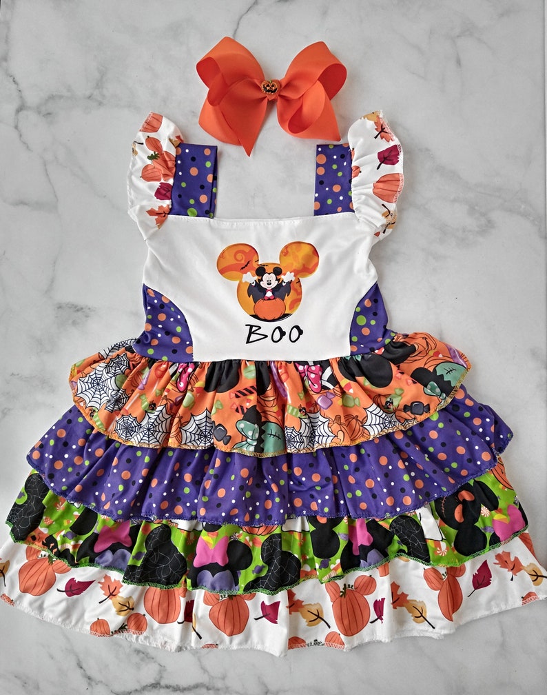 May include: A Halloween dress with a tiered skirt in orange, purple, and green, decorated with pumpkins and leaves. The bodice displays a Mickey Mouse graphic and the word "BOO". An orange bow adds a festive touch.