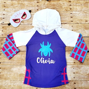 May include: Purple hooded sweatshirt with a spider design and the name "Olivia" on the front. The sleeves have a pink and blue checkered pattern. A white mask with black stitching is included.