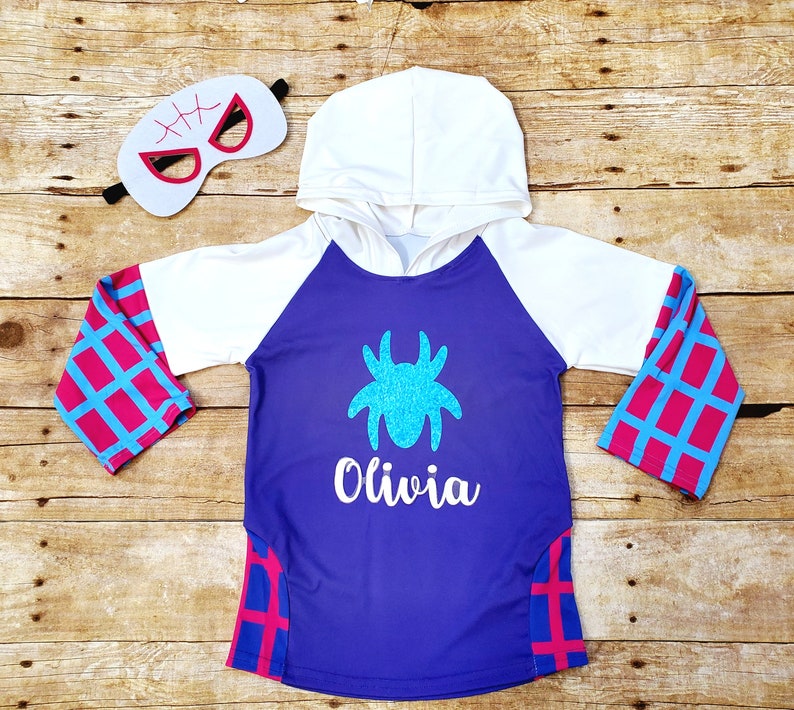 May include: Purple hooded sweatshirt with a spider design and the name "Olivia" on the front. The sleeves have a pink and blue checkered pattern. A white mask with black stitching is included.
