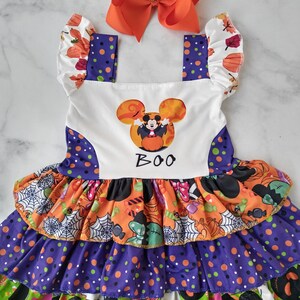 May include: A Halloween dress with an orange bow. The dress has a white bodice with a Mickey Mouse graphic and the word "BOO". The skirt features layers of orange, purple, and green fabric with Halloween patterns.