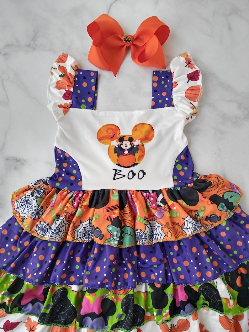 May include: A Halloween dress with an orange bow. The dress has a white bodice with a Mickey Mouse graphic and the word "BOO". The skirt features layers of orange, purple, and green fabric with Halloween patterns.