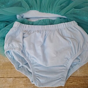 May include: A light blue baby diaper cover with elastic waist and leg openings. A turquoise tulle skirt with sequins is partially visible in the background. The diaper cover is made of soft fabric.