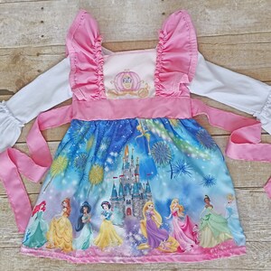May include: A pink and blue princess dress with a white long-sleeved shirt. The dress features a print of Cinderella's castle, fireworks, and all the Disney princesses. The dress has a pink ribbon tie and ruffle sleeves.