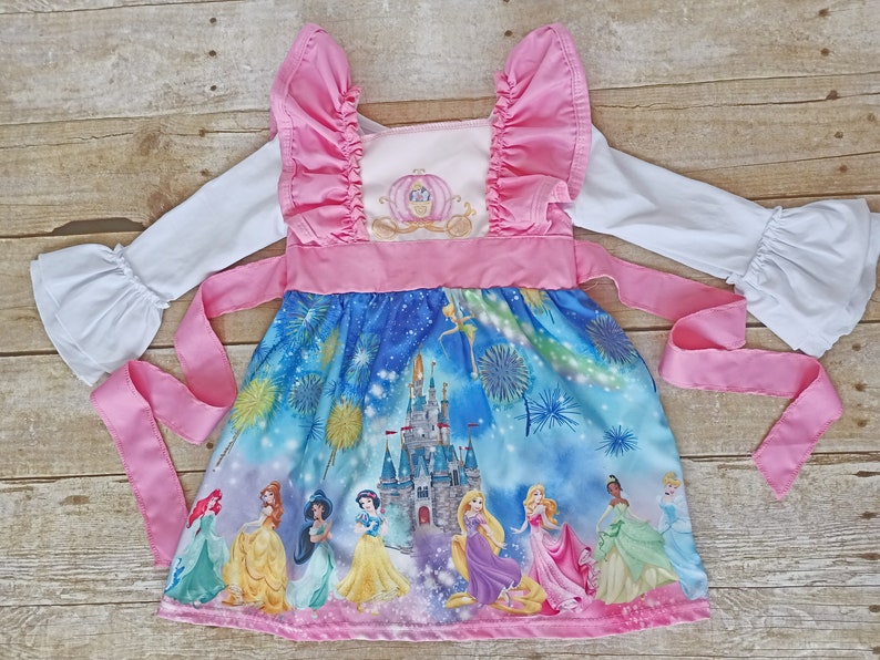 May include: A pink and blue princess dress with a white long-sleeved shirt. The dress features a print of Cinderella's castle, fireworks, and all the Disney princesses. The dress has a pink ribbon tie and ruffle sleeves.