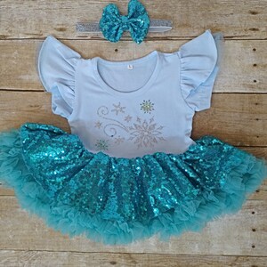 May include: A light blue outfit for a child, featuring a short-sleeved top with a snowflake design and a tutu skirt with sequins. A matching bow and headband complete the set. The outfit is designed for a child.