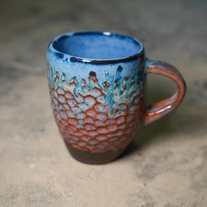 Könnte beinhalten: Eine handgefertigte Keramik-Tasse mit einem strukturierten, wabenartigen Muster. Die Tasse hat einen Farbverlauf von Blau, Türkis und Rotbraun, mit einem dunkelbraunen Boden und Henkel. Der Innenraum ist einfarbig blau.