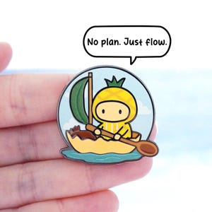 May include: An enamel pin depicting a cartoon pineapple character in a yellow boat with a green sail. The pin includes the text "No plan. Just flow." in a speech bubble, set against a blue sky background.
