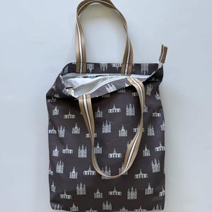May include: A brown tote bag with a repeating white architectural pattern. The bag has tan and white striped handles and a zipper closure. The interior is white. The bag is open, revealing the interior.
