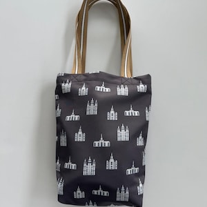 May include: A dark gray tote bag featuring a repeating pattern of white architectural illustrations. The bag has tan and white striped handles. The bag is made of a durable material and is suitable for carrying various items.
