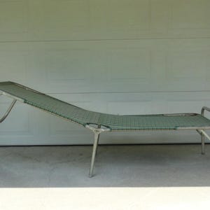 May include: A folding lounge chair featuring a silver-toned metal frame and a green woven mesh fabric seat. The chair has an adjustable backrest section that is currently positioned at an angle, supported by a rear metal frame component.