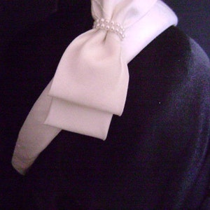 Pu&ograve; includere: Un fiocco di raso color crema con un dettaglio di perle, attaccato a un colletto bianco. Il fiocco &egrave; posto su uno sfondo scuro. Il design a strati del fiocco aggiunge un tocco di eleganza.