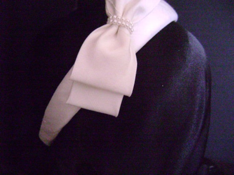 Pu&ograve; includere: Un fiocco di raso color crema con un dettaglio di perle, attaccato a un colletto bianco. Il fiocco &egrave; posto su uno sfondo scuro. Il design a strati del fiocco aggiunge un tocco di eleganza.