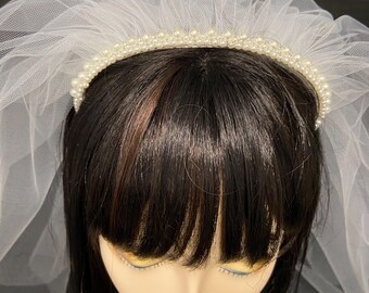 May include: A bridal veil featuring a white tulle design and a pearl-adorned headband. The veil is layered and attached to a crown of small, round pearls. The mannequin head has dark hair.