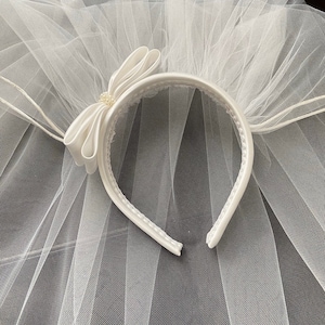 Pu&ograve; includere: Un velo da sposa bianco con una fascia in raso. La fascia presenta un fiocco in raso con un accento di perla. Il velo &egrave; realizzato in un tessuto trasparente a rete.