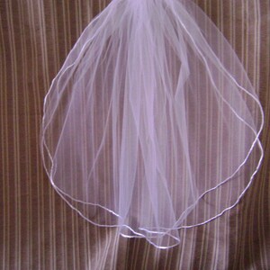 Pu&ograve; includere: Un velo da sposa bianco con bordo smerlato, rifinito con un sottile nastro color argento. Il velo &egrave; realizzato in un tessuto trasparente e leggero ed &egrave; esposto su uno sfondo a righe marroni e bianche.
