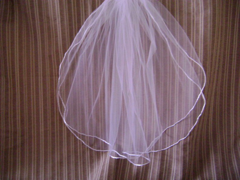Pu&ograve; includere: Un velo da sposa bianco con bordo smerlato, rifinito con un sottile nastro color argento. Il velo &egrave; realizzato in un tessuto trasparente e leggero ed &egrave; esposto su uno sfondo a righe marroni e bianche.