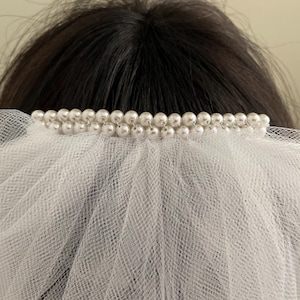 May include: A white bridal veil with a double row of faux pearls along the top edge. The veil is made of a sheer, delicate fabric, and is draped over dark hair.
