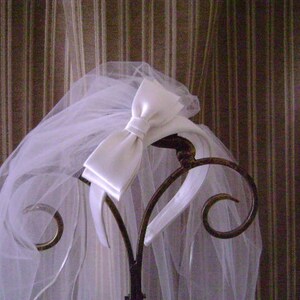 Pu&ograve; includere: Un velo da sposa bianco con un fiocco di raso &egrave; drappeggiato su un supporto decorativo in metallo. Il velo trasparente &egrave; a strati e scorre verso il basso, creando un aspetto morbido e romantico. Il supporto presenta dettagli ornati e vorticosi.