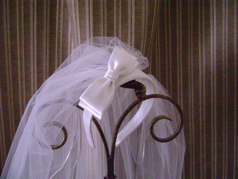 Pu&ograve; includere: Un velo da sposa bianco con un fiocco di raso &egrave; drappeggiato su un supporto decorativo in metallo. Il velo trasparente &egrave; a strati e scorre verso il basso, creando un aspetto morbido e romantico. Il supporto presenta dettagli ornati e vorticosi.