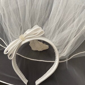 Pu&ograve; includere: Un velo da sposa bianco con una fascia in raso. La fascia presenta un fiocco in raso con un gruppo di perle. Il velo &egrave; realizzato in un tessuto trasparente a rete. La fascia &egrave; larga circa 2.5 cm.