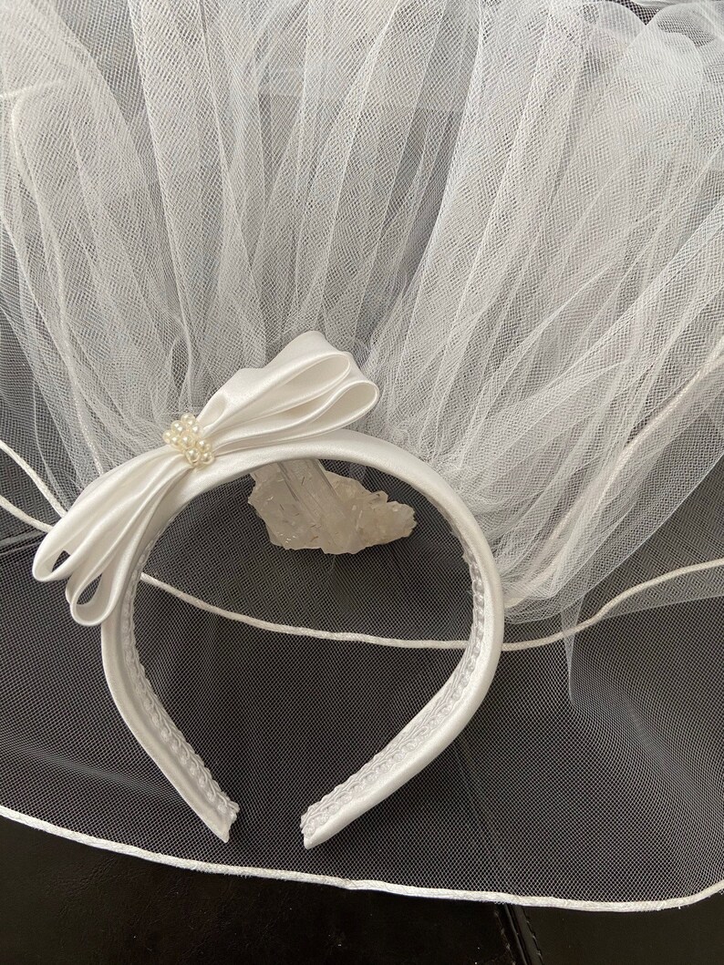 Pu&ograve; includere: Un velo da sposa bianco con una fascia in raso. La fascia presenta un fiocco in raso con un gruppo di perle. Il velo &egrave; realizzato in un tessuto trasparente a rete. La fascia &egrave; larga circa 2.5 cm.