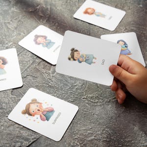 May include: A set of emotion flashcards with illustrations of children expressing feelings like scared, worried, and sad. One card is held by a hand, displaying the word "SAD". The cards are on a gray surface.