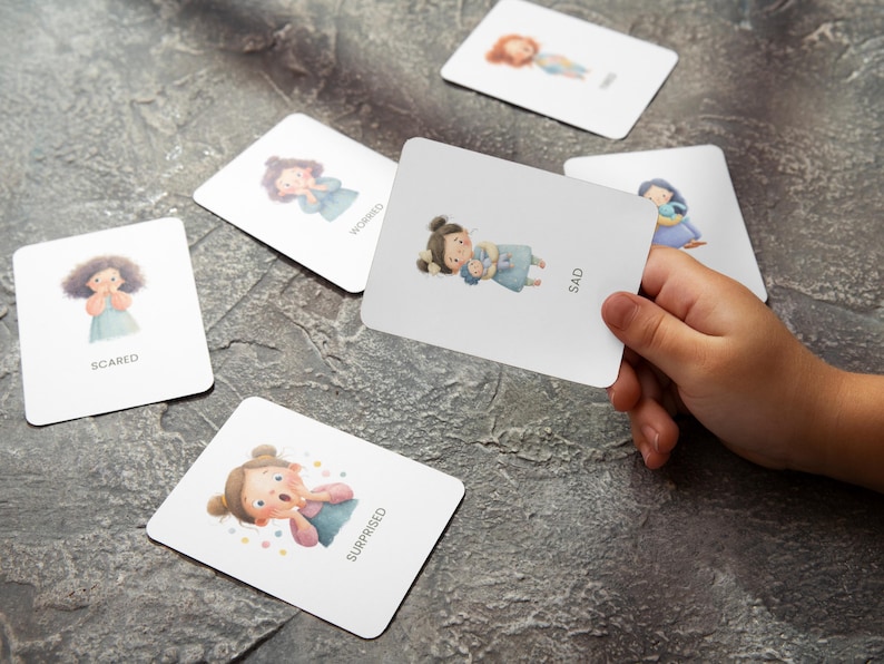 May include: A set of emotion flashcards with illustrations of children expressing feelings like scared, worried, and sad. One card is held by a hand, displaying the word "SAD". The cards are on a gray surface.